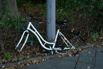 Stripped white bike frame locked to a lamppost, missing wheels, abandoned on a leaf-covered urban pavement. electric bikes mistakes to avoid