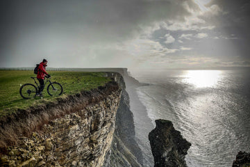 Cyclist on a cliff edge overlooking a dramatic coastal view in the UK at sunset.