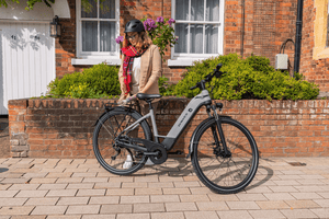 Woman in a helmet standing next to a grey step through electric bike outside a brick house, dressed in a stylish outfit and scarf, ready for an eco-friendly ride.