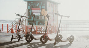 Three electric scooters are parked on a sunlit beachfront promenade with a colourful lifeguard station in the background. 