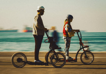 A man and a woman riding electric scooters along a coastal pathway, both wearing helmets, enjoying a smooth and stylish urban commute by the sea.