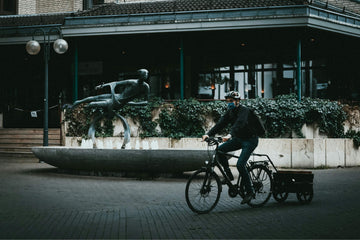 Cyclist with helmet rides an e-cargo bike and trailer through a modern European city square near public art.