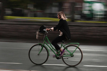 Woman commuting on a green city electric bike with a basket, riding fast through a rainy urban street in smart-casual attire, cycle to work scheme