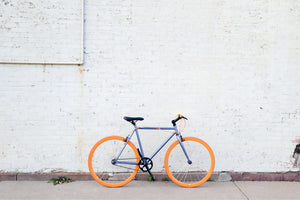 Minimalist fixie bike with bright orange wheels parked against a white urban brick wall. How to choose the best bike for you?