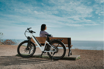 Woman resting on a bench with a white step through frame electric bike beside her, overlooking the ocean under a blue sky.