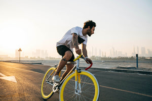 Tattooed cyclist riding a yellow fixie bike on open road at sunrise with city skyline in the background. Are electric bikes worth it?