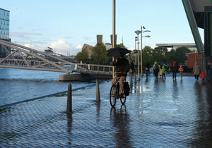 A cyclist riding an electric bike in wet conditions while holding an umbrella, demonstrating all-weather riding capabilities with waterproof and durable design for daily commuting.
