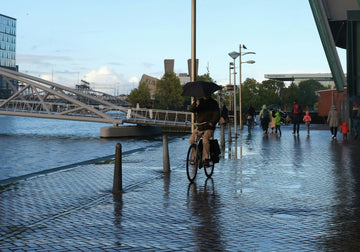 A cyclist riding an electric bike in wet conditions while holding an umbrella, demonstrating all-weather riding capabilities with waterproof and durable design for daily commuting.