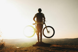 Silhouette of a cyclist lifting an electric hybrid bike at sunrise on a hilltop, symbolising versatility and freedom in urban and off-road environments.