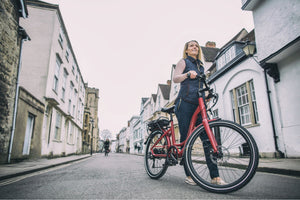 Woman smiling while walking her red Wisper Traditional electric bike through a picturesque town street, combining style and practicality.