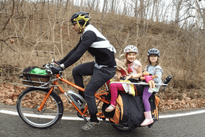 Man riding an electric cargo bike with two children on the back, enjoying a family cycling adventure on a wooded road, highlighting sustainable family transportation.