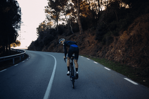 Road cyclist wearing a helmet and sunglasses riding uphill on a winding mountain road, surrounded by trees and rugged terrain, glancing back while cycling in early morning light.