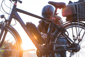 Man in a helmet attaching or removing the battery from an electric bike, with sunlight highlighting the bike frame and a basket on the rear, showcasing the practical setup of an e-bike.