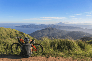 Mountain biker sitting on a grassy hilltop with an orange bike, enjoying a panoramic view of lush green mountains, a serene lake, and distant volcano under a clear blue sky.