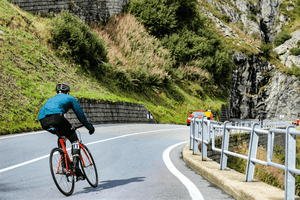 Cyclist in a blue jacket riding up a winding mountain road with steep cliffs, metal guardrails, and a car in the background, navigating a sharp turn surrounded by lush greenery and rocky landscape.