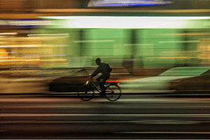 Blurred motion shot of a cyclist riding through a city street at night, with bright lights and passing cars creating a dynamic urban backdrop, emphasising speed and movement.