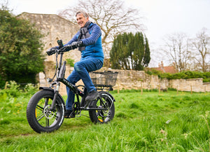 A smiling man rides a black Hygge fat tyre electric bike with a rear basket through a scenic countryside setting. Designed for both adventure and relaxed commuting, this powerful e-bike offers stability, comfort, and long-range performance. Available at Glide & Gear.