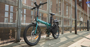 A Hygge folding electric bike in teal, parked in an urban setting with a helmet and pannier bag attached. Designed for city commuting, this compact e-bike features disc brakes, a sturdy frame, and a sleek folding mechanism for easy transport. Available at Glide & Gear.