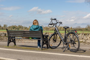A woman in a teal jacket sits on a bench, enjoying the view with a Hygge step-through electric bike parked beside her. Featuring a sleek design, rear rack, and ergonomic saddle, this e-bike is perfect for relaxed rides and daily commuting. Available at Glide & Gear.