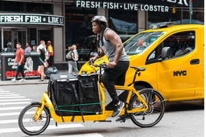 Delivery cyclist on a yellow electric cargo bike navigating busy New York City streets, showcasing urban bike delivery and sustainable transportation.