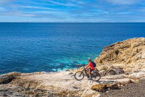 Cyclist riding an electric hybrid bike along a rocky coastline, enjoying a scenic ocean view, highlighting the bike's versatility for adventure and exploration.