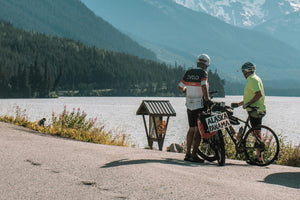 Two cyclists with electric touring mountain bikes taking a break by a scenic lake surrounded by mountains, highlighting the endurance and adventure of long-distance e-bike travel.